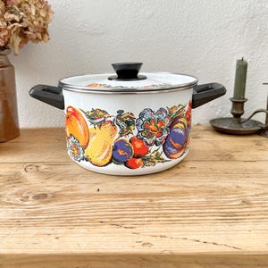 May include: White enamel cooking pot with a black handle and lid. The pot is decorated with a colourful floral and fruit design, including oranges, lemons, and plums. The pot sits on a wooden surface.