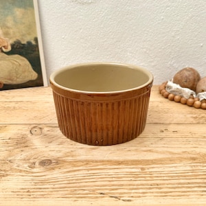 May include: A brown, ribbed ceramic ramekin with a light tan interior. The small, round dish sits on a light-colored wooden surface. A vintage print and decorative items are in the background.