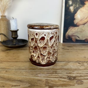 May include: A brown and white ceramic jar with a lid. The jar has a mottled glaze with brown and white swirls. The jar is sitting on a wooden table.