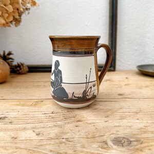 May include: A ceramic mug with a brown rim and handle, featuring a black and white illustration of a person sitting near reeds. The mug has a cream-colored band with the illustration, and a brown base. The mug is on a wooden surface.