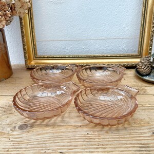 May include: Four vintage, pink glass dishes with a shell-like design. Each dish has a small handle and sits on a wooden surface. A gold-framed picture and other antique items are in the background.