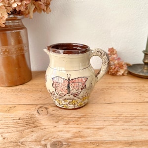 May include: A small, cream-colored ceramic pitcher with a brown rim and handle. The pitcher features a hand-painted butterfly design in pink and red, with yellow floral accents at the base. The pitcher is displayed on a wooden surface.