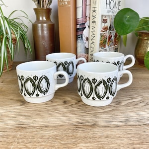 May include: Four white ceramic mugs with a green and white abstract design. The mugs have a rounded shape and a handle on the side. The mugs are sitting on a wooden surface.