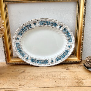 May include: An oval, white serving platter with a teal and gold floral border. The platter is displayed in front of a gold-colored picture frame, resting on a wooden surface. The platter has a scalloped edge.