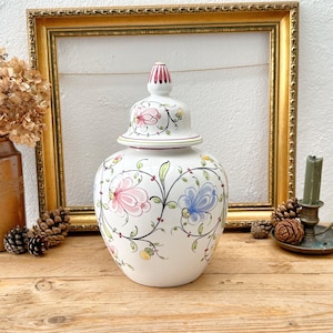 May include: A white ceramic jar with a lid, decorated with pink and blue floral patterns and green vines. The jar is on a wooden surface, with a gold-framed picture behind it. The jar is a decorative item.
