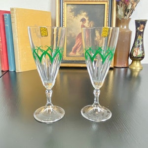 May include: Two clear glass champagne flutes with green and gold designs. The flutes are on a black surface.
