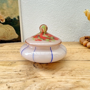 May include: A decorative glass jar with a lid. The jar is a soft pink color with blue vertical stripes and a lid adorned with red and green floral designs. The lid has a small, round, green and orange knob. The jar sits on a wooden surface.