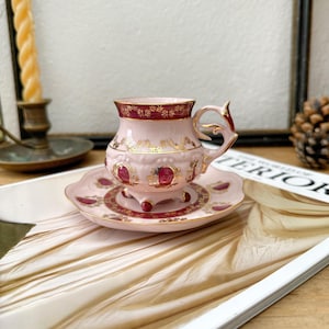 May include: A pink teacup and saucer set with gold detailing. The teacup features an ornate handle and a burgundy rim. The saucer has a complementary design. The set is displayed on a magazine.