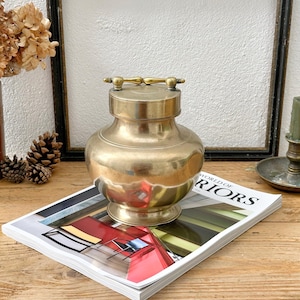 May include: A vintage brass container with a lid and handle, sitting on a magazine. The container has a rounded body and a small base. The magazine has a colorful cover. The background includes a framed picture and dried flowers.