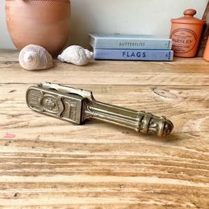 May include: A vintage brass nutcracker with intricate details and a decorative design. The nutcracker is lying on a wooden surface.