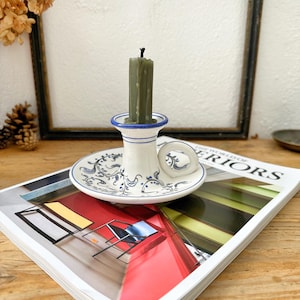 May include: A white ceramic candle holder with blue floral patterns and a green candle. The candle holder rests on a magazine with the text "WORLD OF INTERIORS". The background features a framed picture and dried flowers.