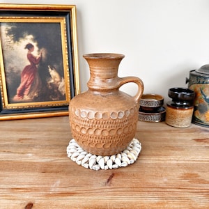 May include: A brown ceramic vase with a textured surface and a handle. The vase is sitting on a white seashell coaster.