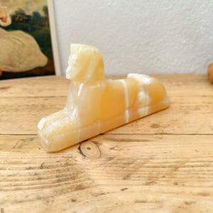 May include: A small, carved figurine of a sphinx in a light amber color. The sculpture has a detailed head and a reclining body, with white veining throughout the stone. The figurine is displayed on a wooden surface.