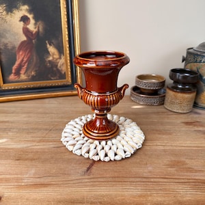May include: A brown ceramic urn vase with a white seashell coaster. The vase has a classic design with a round base and a flared top.