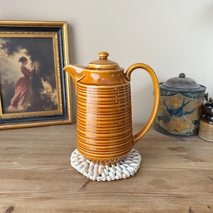 May include: An antique, amber-glazed ceramic teapot with a ribbed pattern, a curved handle, and a lid. The teapot is placed on a white woven coaster. A framed painting and other vintage items are in the background.