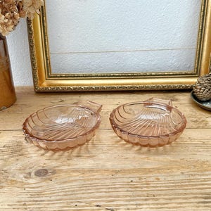 May include: Two pink glass seashell-shaped dishes with ridged detailing. The dishes are on a wooden surface, with a gold-framed picture and dried flowers in the background. These decorative dishes are ideal for serving small portions.