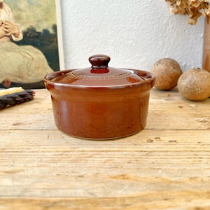 May include: A small, round, brown ceramic casserole dish with a lid. The dish has a handle on top of the lid and is sitting on a wooden surface. The dish is likely used for cooking and serving food.