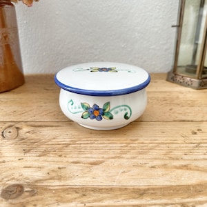 May include: A small, white ceramic trinket box with a lid. The box is adorned with blue and green floral designs and a blue rim. The lid also features a floral design. The box sits on a wooden surface.