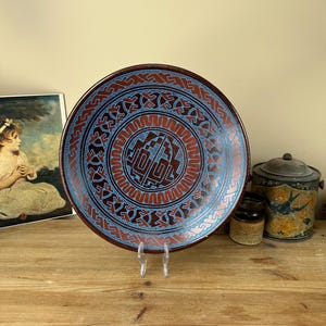 May include: A large, round ceramic plate with a brown glaze and intricate blue and red geometric designs. The plate has a raised rim and a central design that resembles a stylised sun or flower.