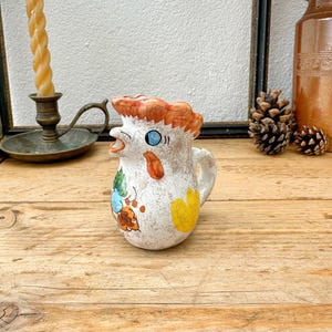 May include: A small, vintage ceramic pitcher with a rooster design. The pitcher is white with orange, blue, and yellow accents. It has a handle and a spout shaped like a rooster's head. The pitcher is on a wooden surface.
