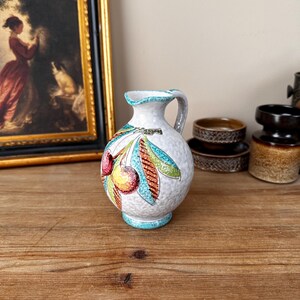 May include: A small white ceramic jug with a blue rim and a handle. The jug is decorated with a colourful cherry design.