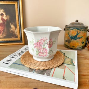 May include: A white ceramic planter with a floral design featuring pink and white flowers. The planter is octagonal in shape and has a slightly flared rim. It is sitting on a woven wicker coaster.
