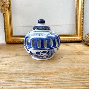 May include: A blue and white ceramic jar with a lid. The jar has a round shape with a series of vertical openings. The design includes floral patterns and decorative swirls. The jar is placed on a wooden surface.