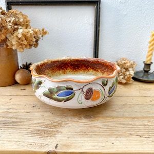 May include: A ceramic bowl with a white base and a scalloped rim. The bowl features hand-painted green, blue, and orange floral designs. The interior has a brown and orange gradient. The bowl sits on a wooden surface.