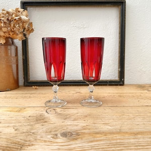 May include: Two tall champagne flutes featuring deep red glass bowls with vertical cut patterns. Each glass has a clear, faceted stem and a circular base, standing upright with a slender, cylindrical shape and a polished, reflective finish.