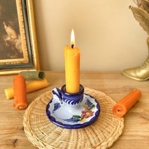 May include: A lit orange candle in a blue and white floral ceramic holder sits on a woven wicker coaster. Several unlit orange and green candles are nearby. The candle holder has a handle.