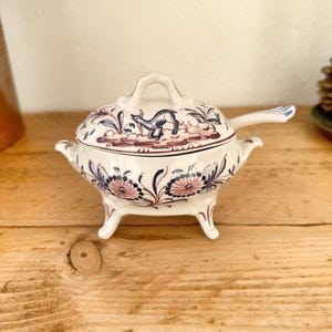 May include: A white ceramic sugar bowl with a lid and spoon, featuring a blue and pink floral design. The bowl has three small feet and a handle on the lid. The lid has a blue animal illustration.