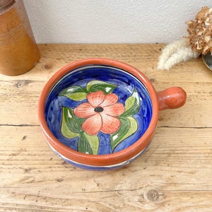 May include: Hand-painted ceramic bowl with a handle. The bowl features a vibrant floral design with a red flower, green leaves, and a blue background. The rim is a reddish-orange colour. A decorative piece for the kitchen.
