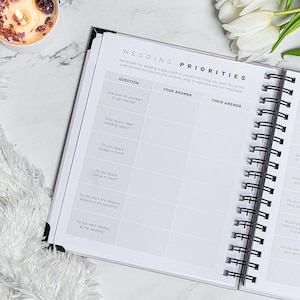 Pu&ograve; includere: Un'agenda per matrimoni a spirale aperta su una pagina intitolata "WEDDING PRIORITIES" con sezioni di domande e risposte. Una candela accesa e fiori bianchi sono sullo sfondo.