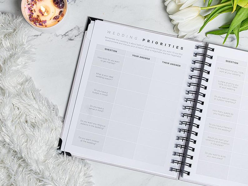 Pu&ograve; includere: Un'agenda per matrimoni a spirale aperta su una pagina intitolata "WEDDING PRIORITIES" con sezioni di domande e risposte. Una candela accesa e fiori bianchi sono sullo sfondo.