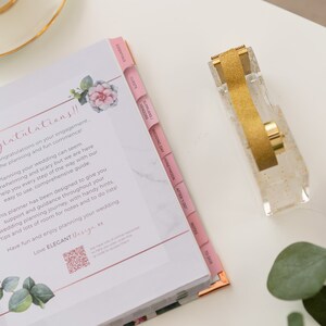 May include: A pink wedding planner with tabs for essentials, guests, vendors, suppliers, wedding day, honeymoon, after-do, notes, and to-dos. The planner has a floral design and a QR code. A gold glitter tape dispenser is on a white surface next to the planner.