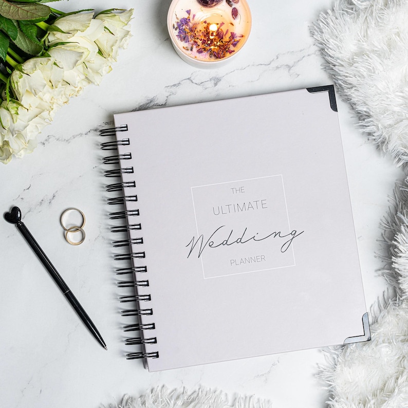 Pu&ograve; includere: Un'agenda per matrimoni a spirale grigio chiaro con la scritta "THE ULTIMATE Wedding PLANNER" sulla copertina. L'immagine include una penna nera con la parte superiore a forma di cuore, anelli d'oro, una candela accesa e rose bianche.