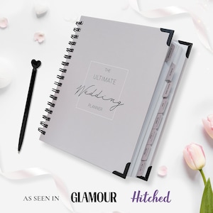 Pu&ograve; includere: Un'agenda per matrimoni grigio chiaro con rilegatura a spirale con il testo "THE ULTIMATE Wedding PLANNER". &Egrave; visibile una penna nera con la parte superiore a forma di cuore e divisori etichettati. Presentato in Glamour e Hitched.