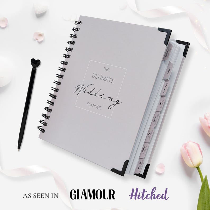 Pu&ograve; includere: Un'agenda per matrimoni grigio chiaro con rilegatura a spirale con il testo "THE ULTIMATE Wedding PLANNER". &Egrave; visibile una penna nera con la parte superiore a forma di cuore e divisori etichettati. Presentato in Glamour e Hitched.