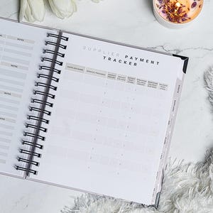 Pu&ograve; includere: Planner a spirale aperto con copertina bianca e rilegatura a spirale nera. Le pagine sono aperte su una pagina "Supplier Payment Tracker" con colonne per il nome del fornitore e i dettagli di pagamento. Una candela e dei fiori sono sullo sfondo.