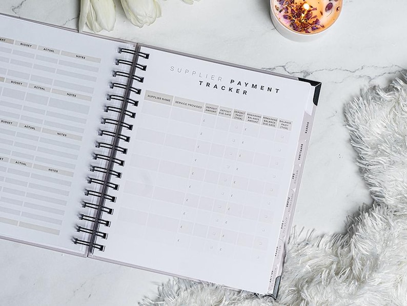 Pu&ograve; includere: Planner a spirale aperto con copertina bianca e rilegatura a spirale nera. Le pagine sono aperte su una pagina "Supplier Payment Tracker" con colonne per il nome del fornitore e i dettagli di pagamento. Una candela e dei fiori sono sullo sfondo.