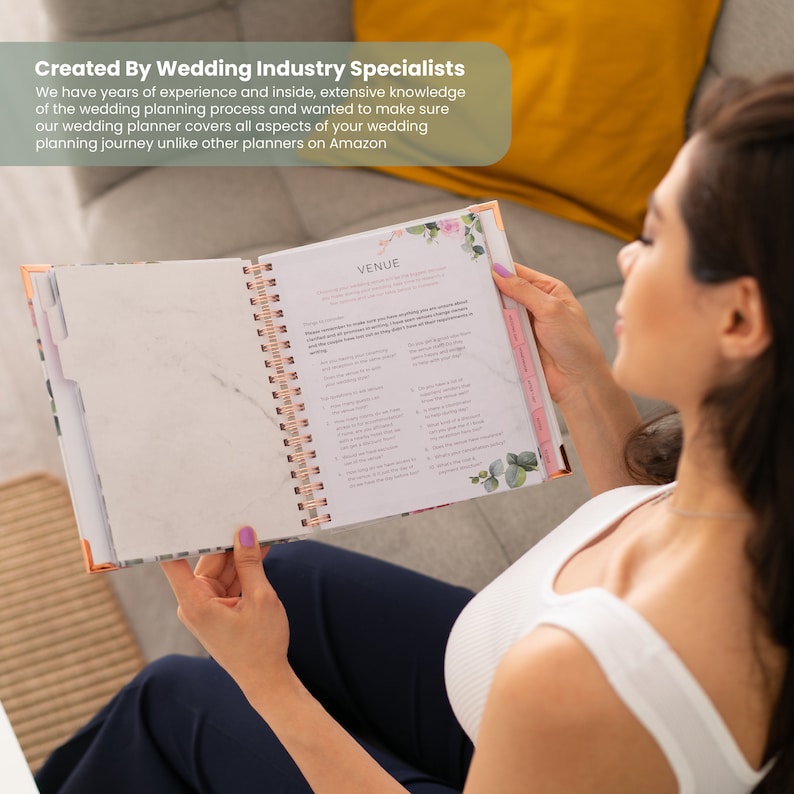 May include: A wedding planner with a floral design on the cover. The planner is open to a page titled "Venue" with a checklist of questions to ask potential venues. The planner is being held by a person.