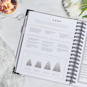 Pu&ograve; includere: Planner per matrimoni aperto con rilegatura a spirale, con sezioni su considerazioni sulla torta e dimensioni delle torte pi&ugrave; diffuse. Il planner &egrave; su una superficie di marmo, con una candela accesa e tulipani bianchi sullo sfondo.