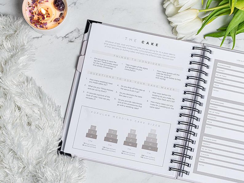 Pu&ograve; includere: Planner per matrimoni aperto con rilegatura a spirale, con sezioni su considerazioni sulla torta e dimensioni delle torte pi&ugrave; diffuse. Il planner &egrave; su una superficie di marmo, con una candela accesa e tulipani bianchi sullo sfondo.