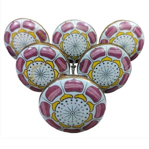 Op de afbeelding: Zes keramische kastknoppen met een witte achtergrond en een roze en geel bloemenontwerp. De knoppen hebben een gouden rand.