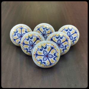 May include: Five ceramic knobs with a white background and a blue and yellow floral pattern. The knobs have a raised edge and a small red dot in the center of the floral design.