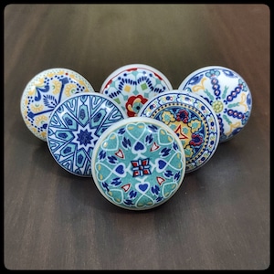 Op de afbeelding: Zes keramische knoppen met ingewikkelde blauwe, groene, gele en rode patronen. De knoppen zijn rond en hebben een witte achtergrond.