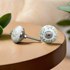 Op de afbeelding: Twee witte keramische kastknoppen met een lichtgroen bloemmotief. De knoppen hebben een zilverkleurige metalen basis en een schroefdraadstang voor installatie. Eén knop ligt, de andere staat rechtop.