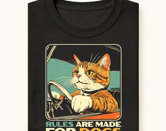 May include: Black t-shirt featuring a cartoon cat driving a car, with orange and white fur and a black collar. The image includes the text "RULES ARE MADE FOR DOGS" in a retro style font. The shirt is folded.