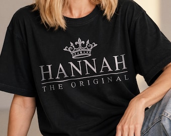 May include: A black short-sleeved t-shirt featuring a silver graphic print. The design includes a crown icon above the text "HANNAH" in large capital letters, with the words "THE ORIGINAL" printed in smaller capital letters directly underneath.