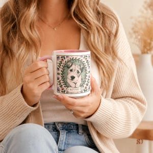 May include: White ceramic mug with a pink handle and rim, featuring a portrait of a dog named "Winnie" surrounded by a floral wreath. The mug is held by a person, showcasing the design and color scheme.
